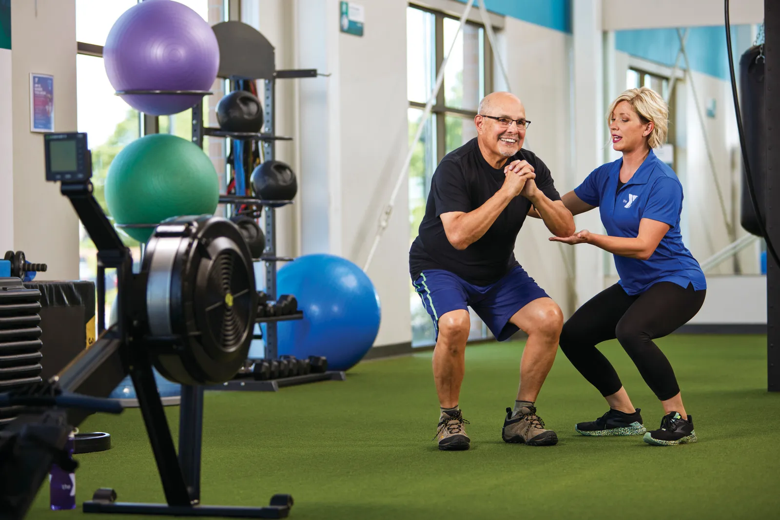 Trainer coaching an older man through a squat in a bright fitness studio