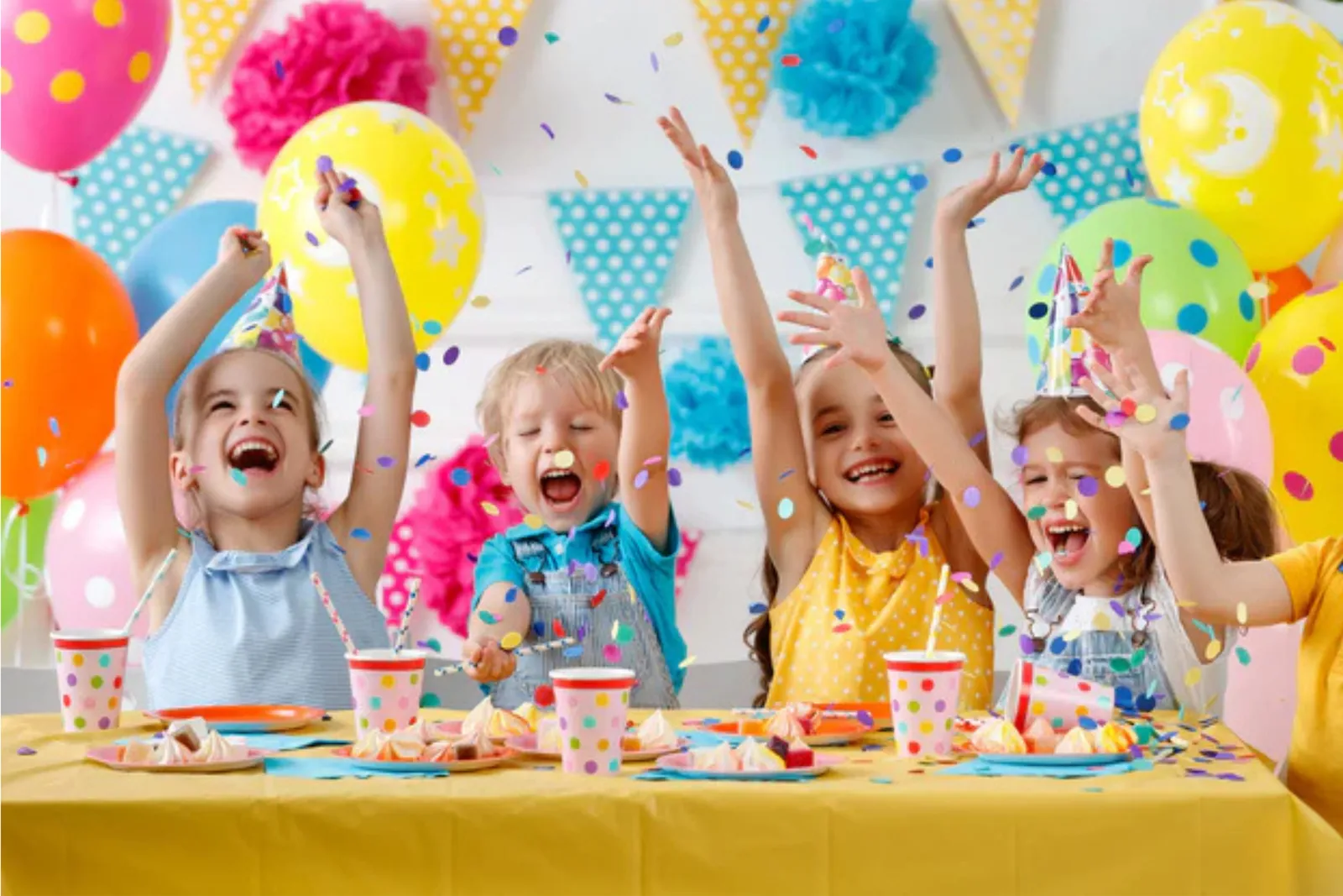 Kids in party hats throwing confetti at a birthday table decorated with balloons and bunting